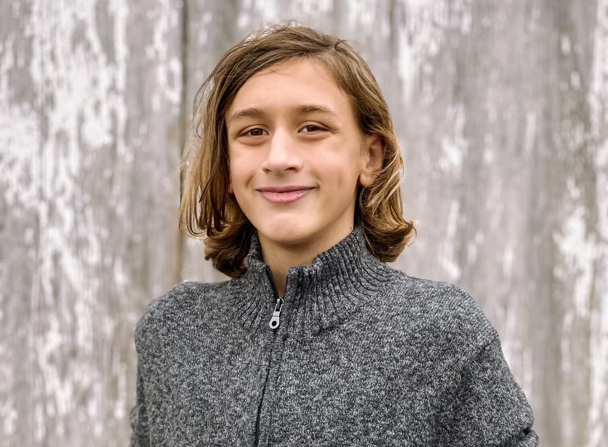 A smiling boy with shoulder-length hair poses in a gray zip sweater against a weathered wood background