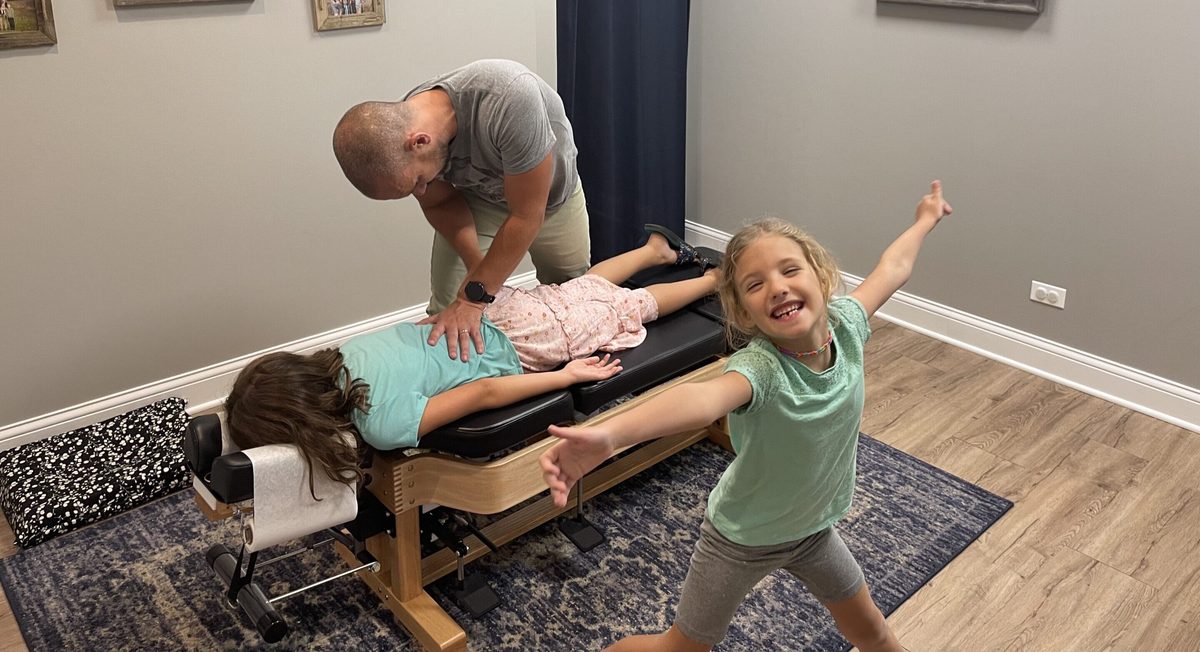 A man adjusts a girl lying on a treatment table while another girl jumps and smiles beside them