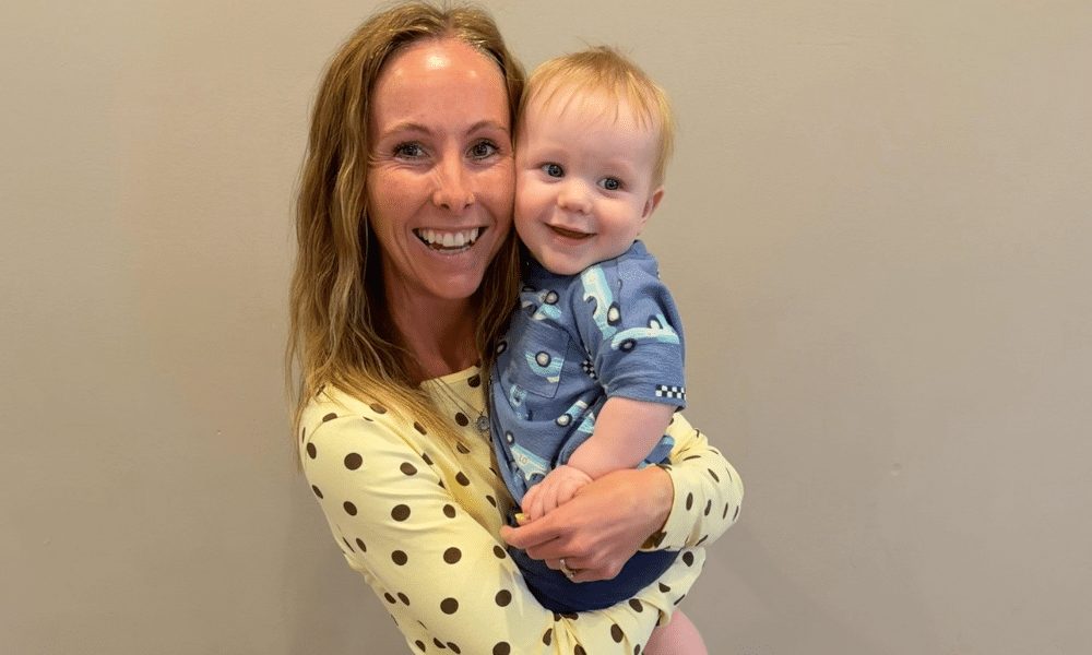 A woman in a polka dot top smiling while holding a baby in a blue patterned outfit against a plain wall