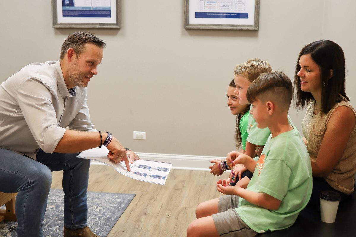 A man points at a paper while three children and a woman sit nearby listening in a clinic hallway
