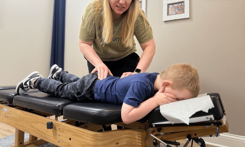 An adult gently adjusts a boy's lower back as he lies face down on a chiropractic table, covering his face with his hands.