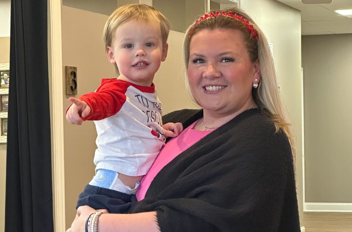 A woman holds a toddler boy who points off to the side in a hallway