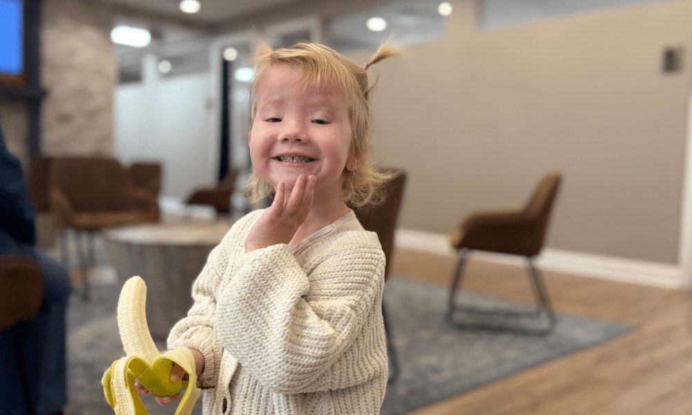 A young girl with pigtails in a chiropractic office lobby holding a peeled banana up near her chin and smiling