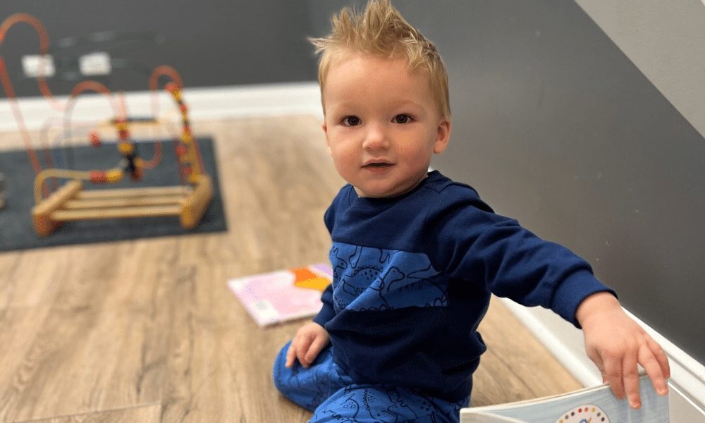 A toddler with spiky blond hair sitting on a wood floor reaching for a board book, with a bead maze toy in the background