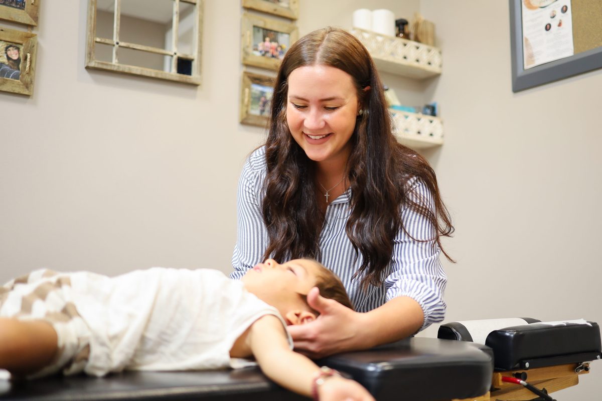 A smiling woman gently touching the neck of a calm child lying face-up on a treatment table