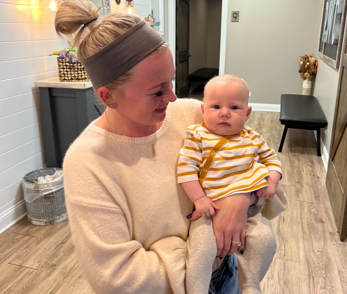 A woman holding a baby in a striped onesie in a home hallway, looking down at the baby