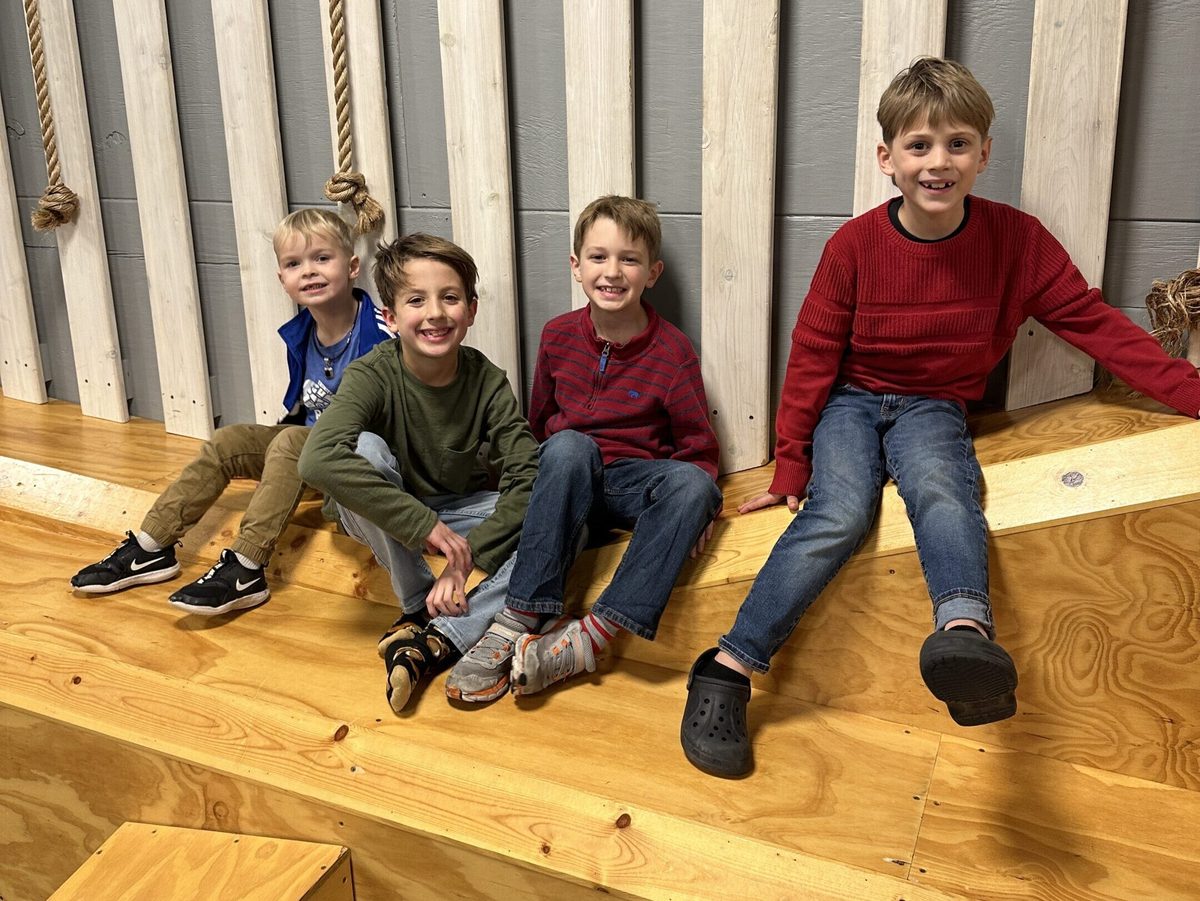 Four young boys sitting together and smiling on a wooden indoor play structure