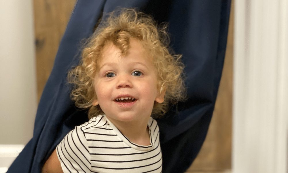 A curly-haired toddler in a striped shirt smiling in front of a navy curtain