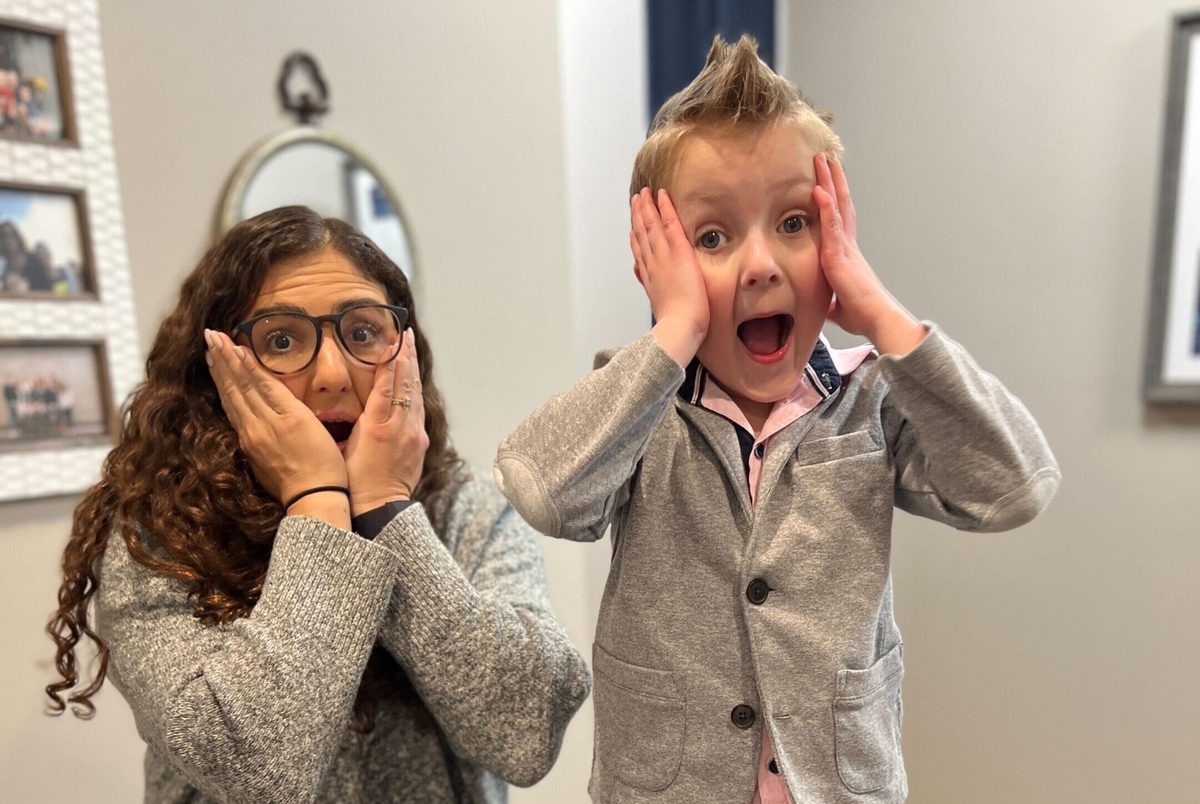 A mom and her young son making surprised, wide eyed faces with their hands pressed to their cheeks in a home hallway