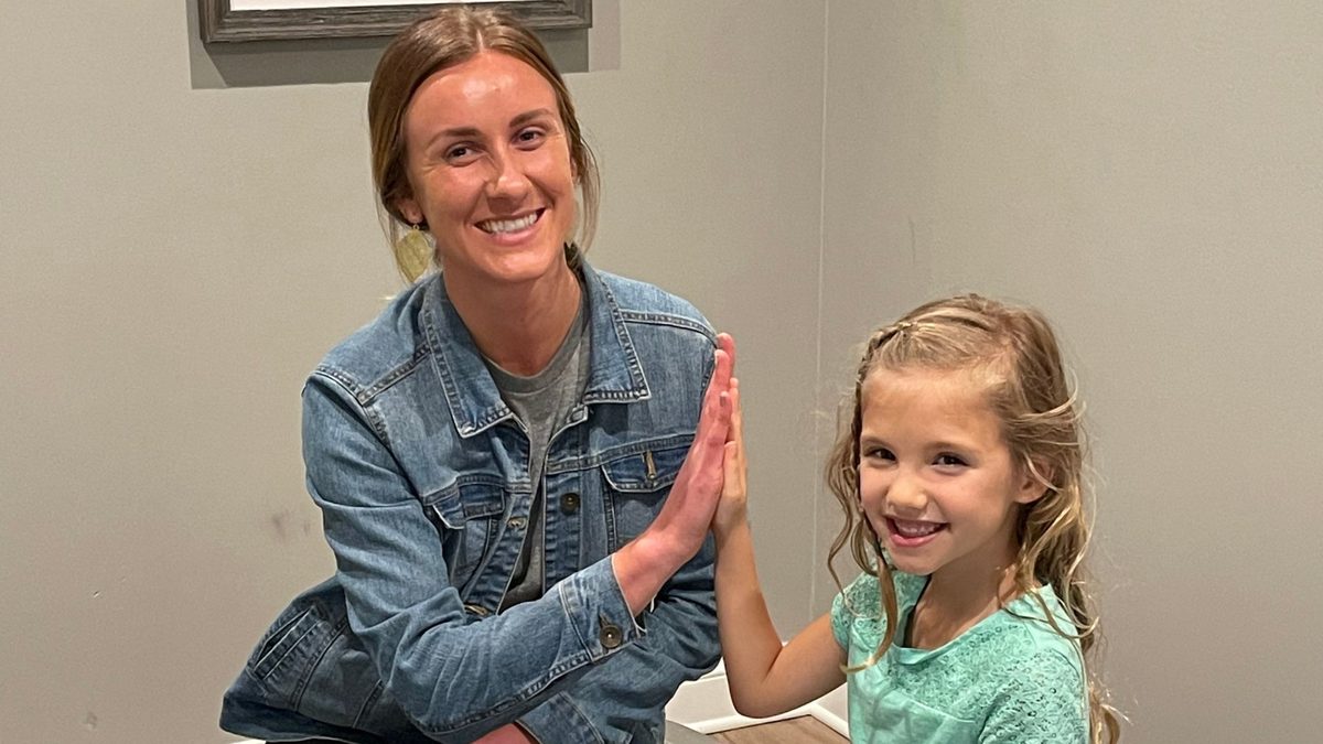 A woman in a denim jacket high-fiving a young girl in a teal shirt, both smiling in a room with a plain gray wall