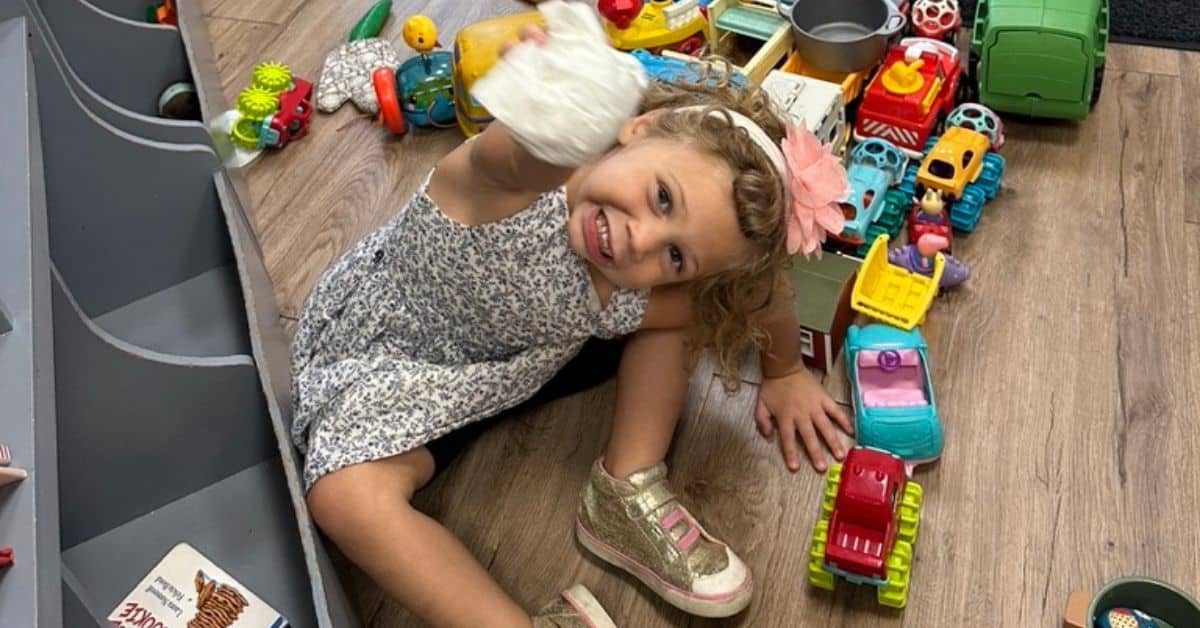 A young girl sitting on the floor amid scattered toys, smiling and holding up a soft white stuffed toy above her head