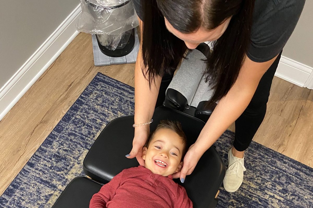A woman gently supporting a smiling toddler's head and neck while the toddler lies on a treatment table