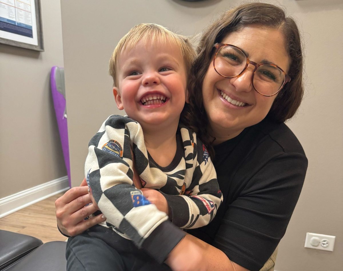 A woman with glasses hugging a laughing toddler boy in a checkered outfit in a chiropractic office lobby