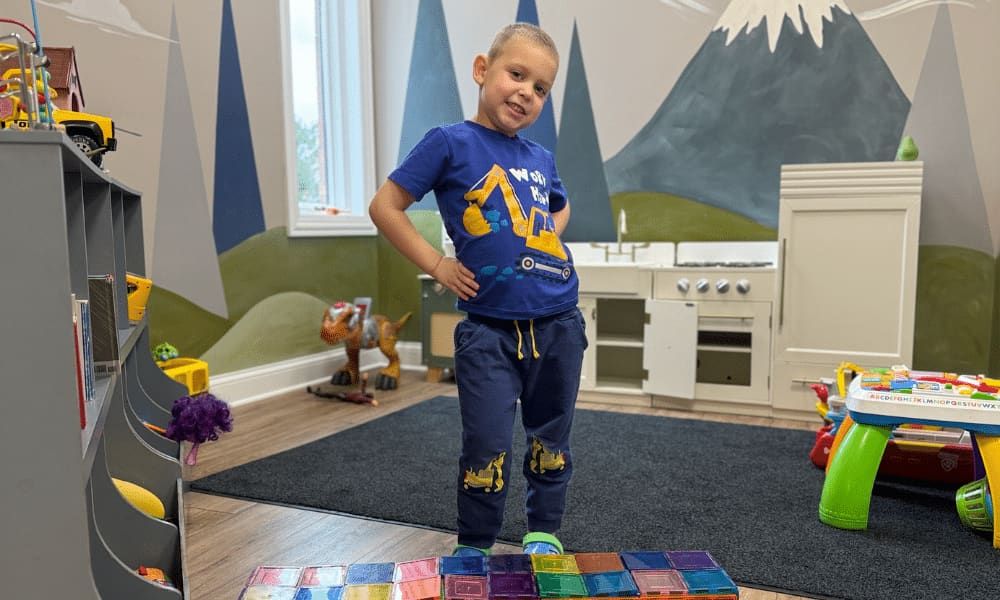 A smiling boy stands in a playroom with a painted mural and toys.