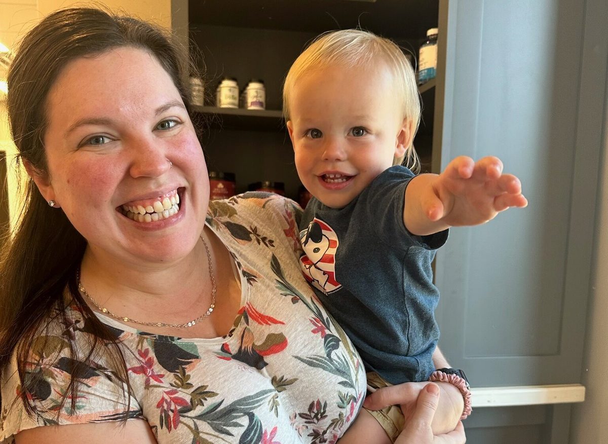 A woman holding a laughing toddler who reaches toward the camera, with supplement bottles on shelves behind them