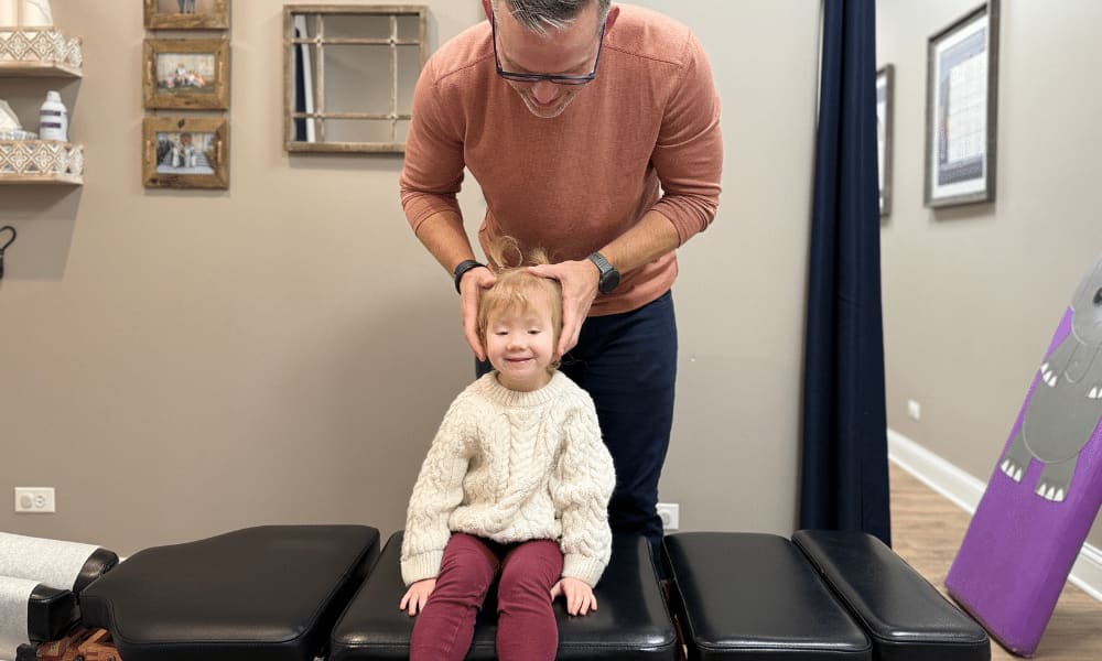 A man in glasses gently holding a smiling young girl's head while she sits on a treatment table