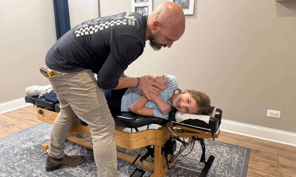 A man gently supports a smiling girl's shoulder and hip as she lies on her side on a chiropractic table.