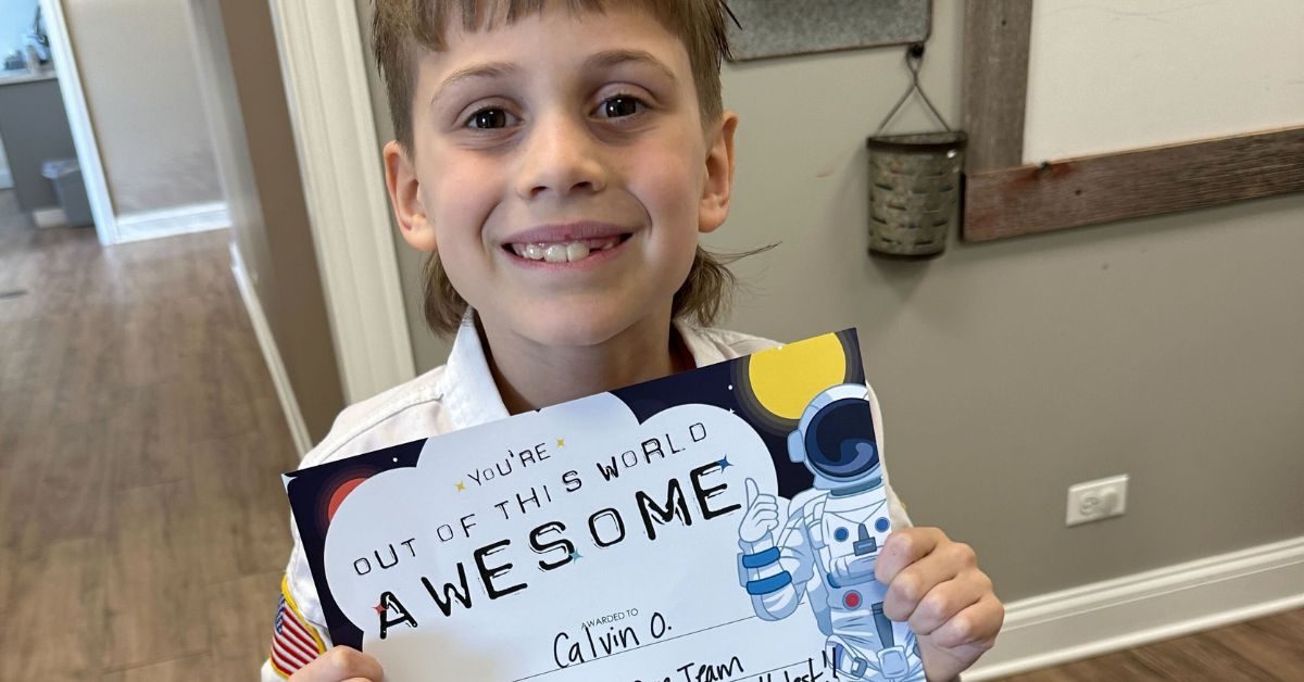 A boy in a martial-arts uniform smiling while holding up an award certificate decorated with an astronaut illustration, standing in a hallway.