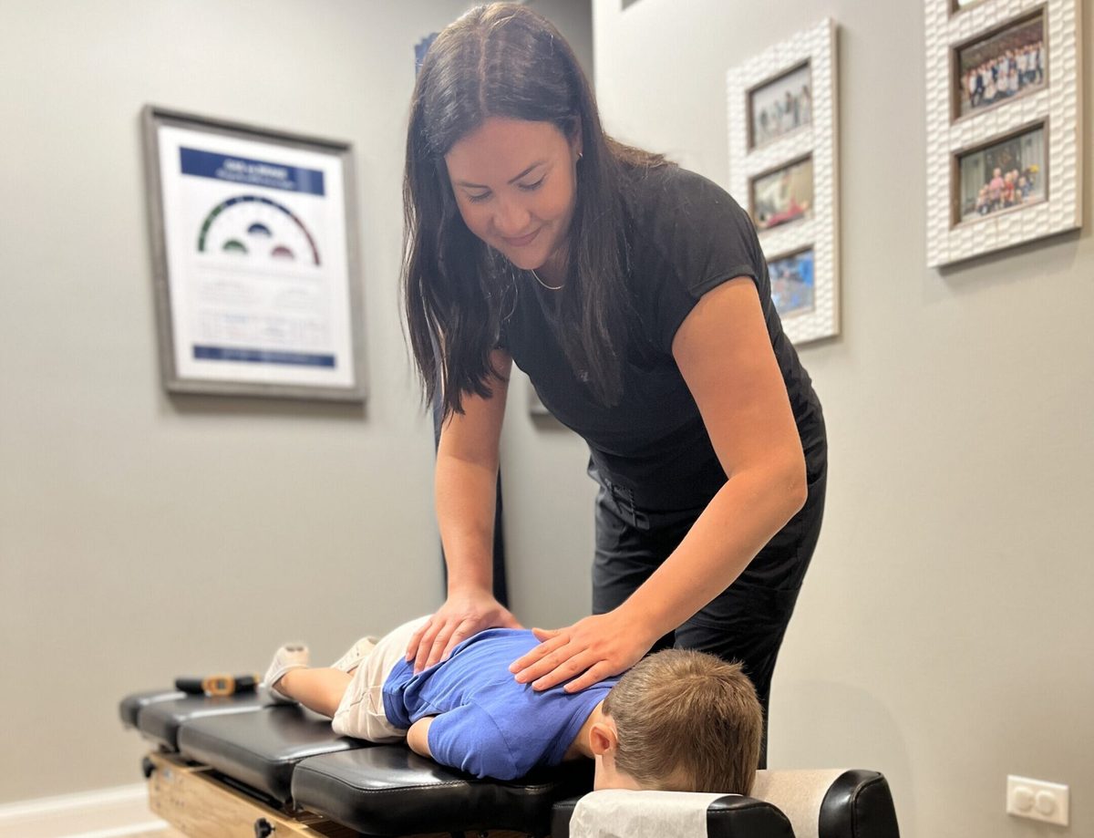 A chiropractor gently pressing on a young boy's mid back while the boy lies face down on a treatment table