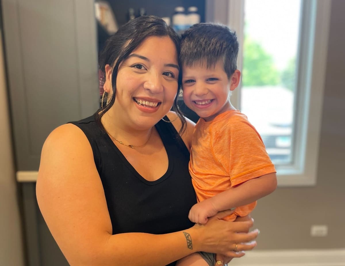 A smiling woman holding a smiling young boy in an orange shirt indoors