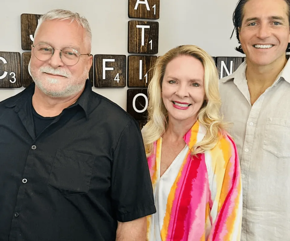 Dr. John Johr, Dr. Anna Saylor and Dr. Zach Conner smiling together in front of the letter tile wall in our office